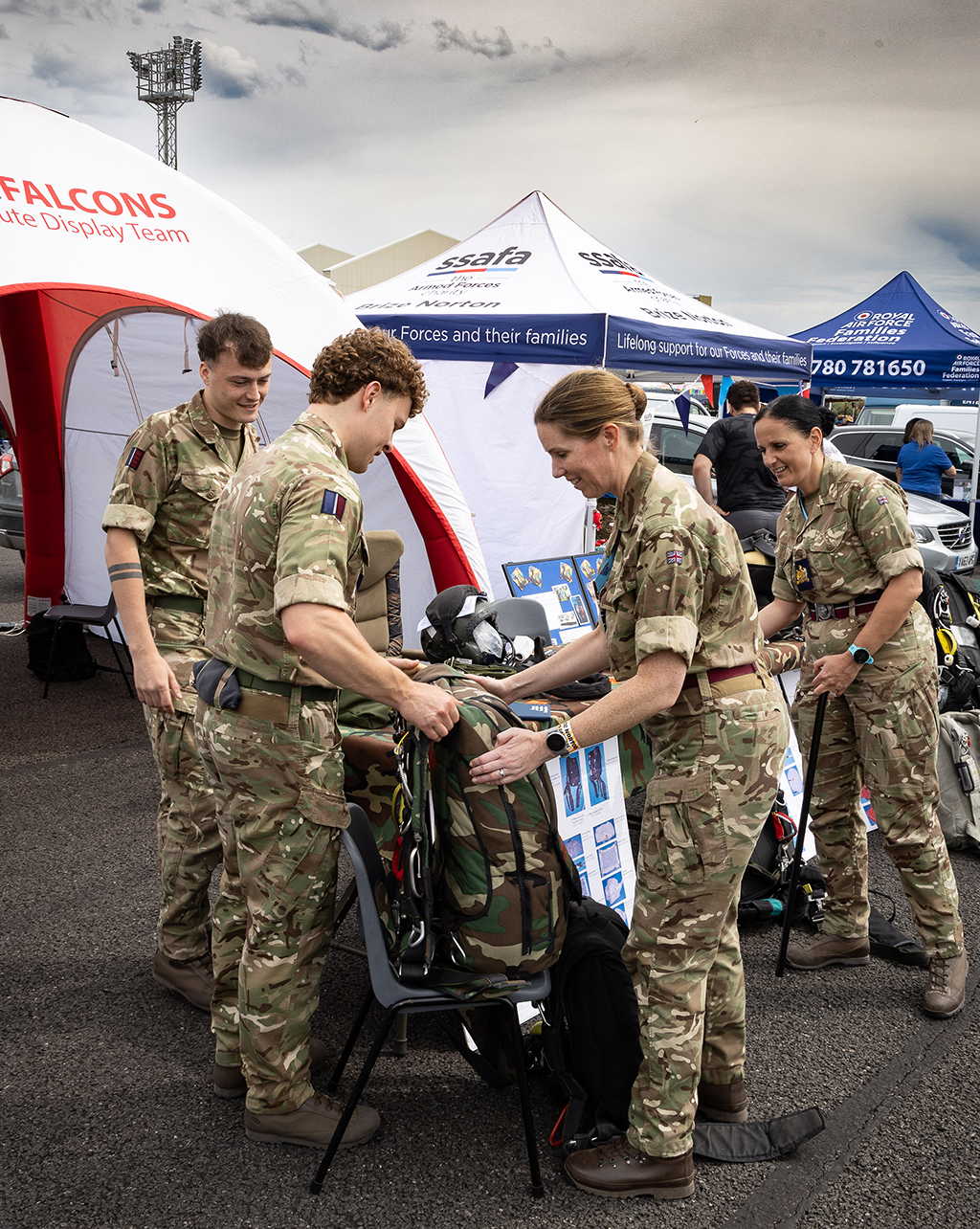 The Station Commander and Station Warrant Officer, wearing camouflage uniforms, interacting with other personnel and a backpack parachute at a display tent, with informational posters and "SSAFA" charity banners in the background